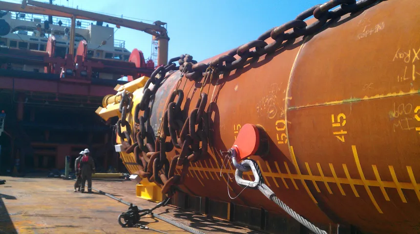 A suction pile, namely a long steel tube, 16 ft in diameter, with 2 men in coveralls next to it on the back deck of a vessel 